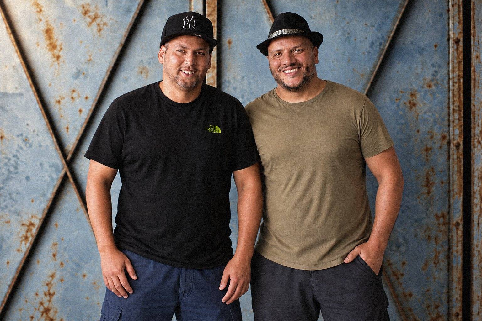 KamC, in the black T-shirt and cap, and RACKS, in the khaki T-shirt and hat, in front of a metal door